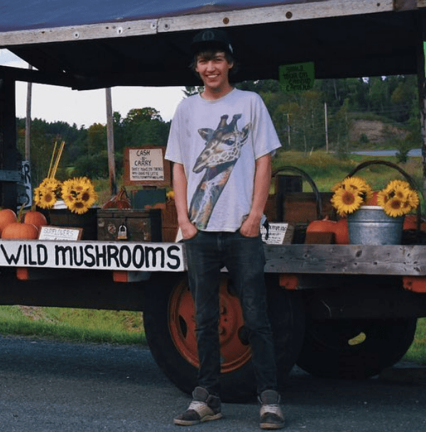 Ben Decker wearing a flat brim hat, jeans and a colorful tshirt with his hands in his pockets smiling in at the camera. He stands in front of a open air carriage that sells local farm products.